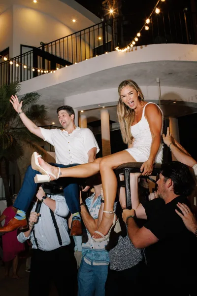 Wedding reception dance as guests lift bride and groom on chairs during the hora, under string lights in an indoor venue with arched ceiling