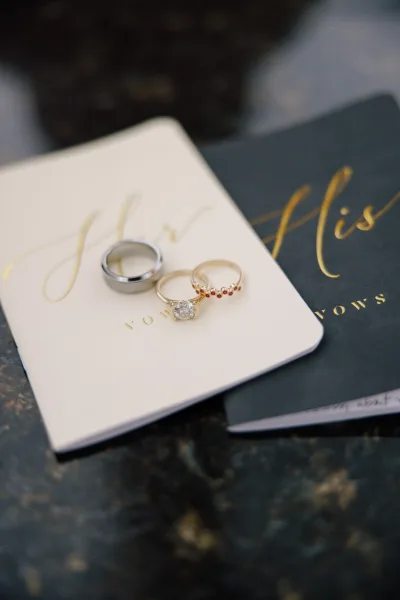 Wedding rings with engagement ring and band resting on black and white vow books, diamond solitaire and ruby ring on a marble surface