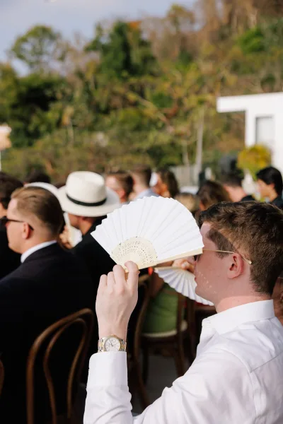 Wedding guests seated at an outdoor ceremony, one man in sunglasses and fedora holding a white folding fan among wooden chairs by trees