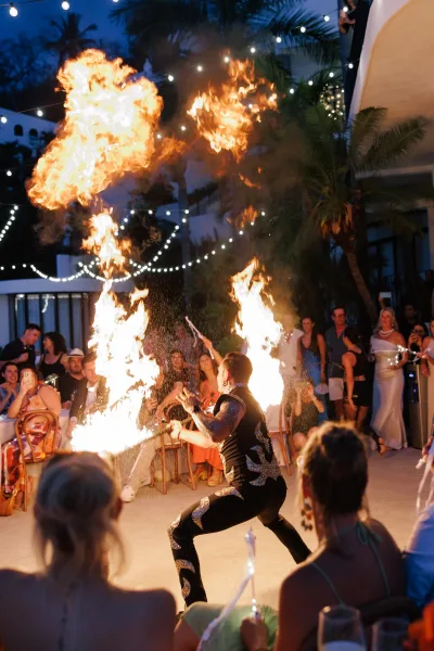 Wedding fire performance with fire torches as guests watch on an outdoor patio under string lights, palm trees, and night sky backdrop