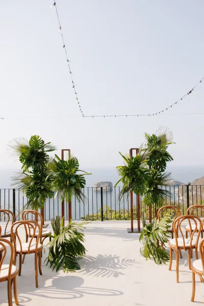 Ceremony setup for an outdoor wedding ceremony with a wooden arch and tropical greenery on a terrace overlooking ocean cliffs under string lights