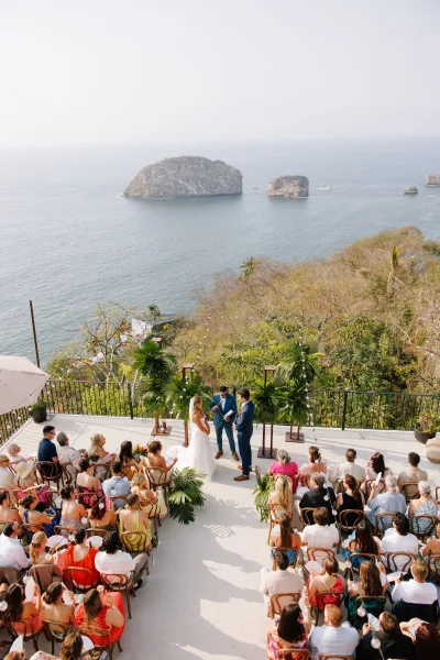 Wedding ceremony with bride and groom under a tropical greenery arch, guests seated on wooden chairs on a cliffside terrace with ocean view