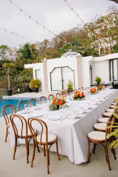 Reception tablescape with a long banquet table wedding setup, white linens and orange floral centerpieces under string lights by a modern villa pool