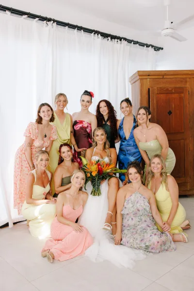 Bridesmaid group photo with bride with bridesmaids in mismatched dresses, holding a tropical bouquet, posed in an indoor room with white curtains