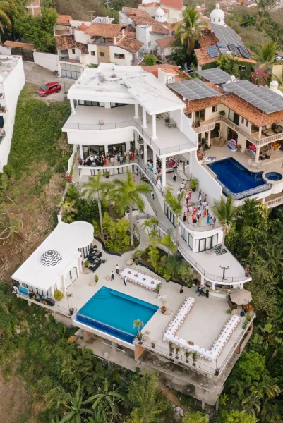 Wedding reception setup with long banquet tables in white linens by a pool, set on a hillside villa terrace with palm trees and umbrellas