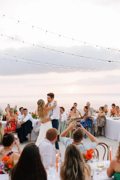 First dance outdoors as bride and groom sway under string lights beside long guest tables, with ocean horizon and sunset sky behind