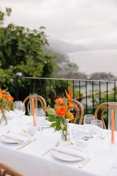 Reception tablescape with an outdoor reception table of white linens, gold flatware, orange taper candles and tropical florals on a coastal terrace overlooking the ocean