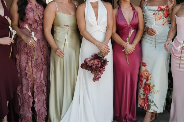 Bridesmaid dresses in a mismatched bridesmaid dresses palette, bridesmaids holding calla lilies with ribbon bows against a neutral wall with greenery