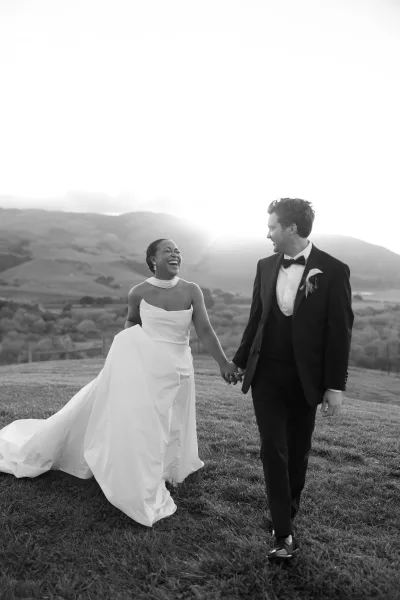 Couple portrait of bride and groom walking hand in hand, bride in veil and train, groom in tux, across a field at sunset mountains