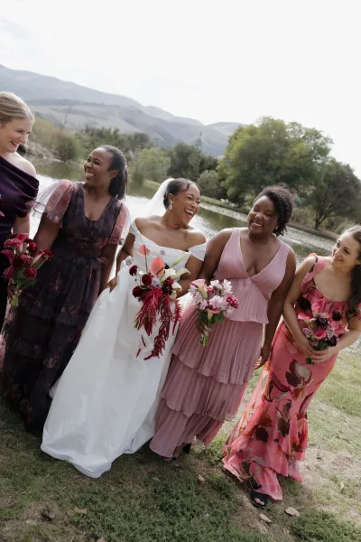 Bridesmaids portrait with bride in veil holding bouquet beside bridesmaids in mismatched dresses on a lakeside lawn with mountains beyond