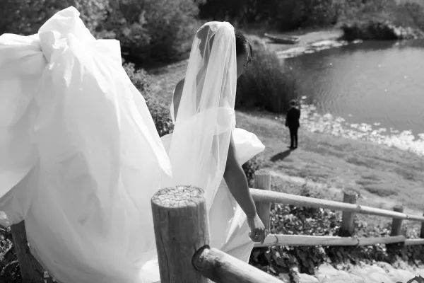 Wedding couple portrait from behind as bride in veil and tulle gown holds her train, approaching groom in suit by a lakeside fence