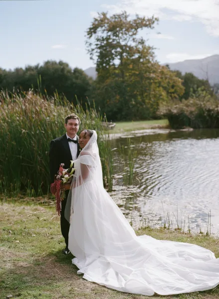Couple portrait of bride holding a calla lily bouquet beside groom in black tuxedo, her veil and long train flowing by a mountain lake