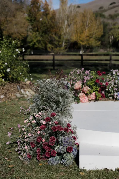 Ceremony aisle decor with floral aisle markers of pink, burgundy, and cream roses, carnations, and hydrangea on a lawn near a white platform