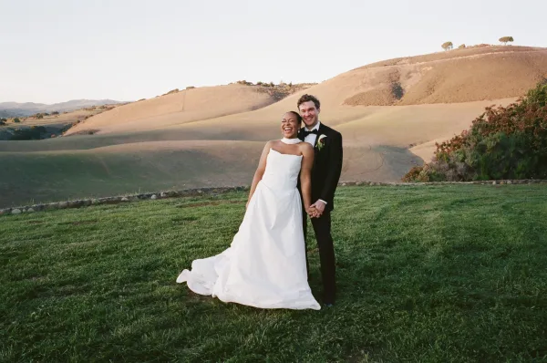 Couple portrait of bride and groom holding hands in a grassy field, bride laughing in strapless dress as groom stands behind her, hills beyond