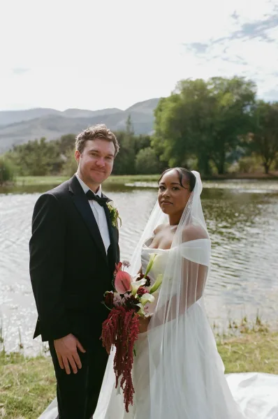 Couple portrait of bride and groom, her off-the-shoulder dress and veil with bouquet, by a lake with mountains and cloudy sky