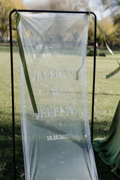Wedding welcome sign with sheer fabric hanging on a black metal frame, green ribbon accent, set on a grassy lawn with trees