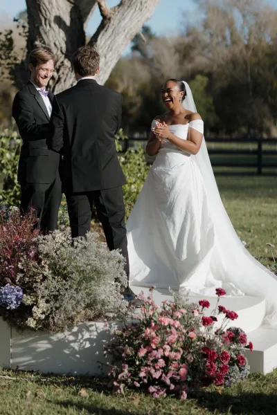 Wedding ceremony moment as bride laughs in strapless dress and long veil facing groom in bow tie on a floral platform under trees and blue sky