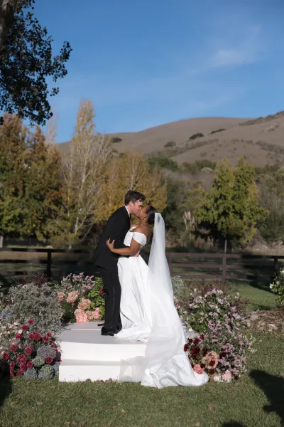 Wedding kiss portrait of bride and groom kissing, her long veil flowing beside pink and burgundy florals on ceremony steps outdoors