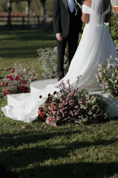 Ceremony moment as bride in off the shoulder wedding dress and veil faces tuxedo groom on a white platform amid garden aisle florals on grass