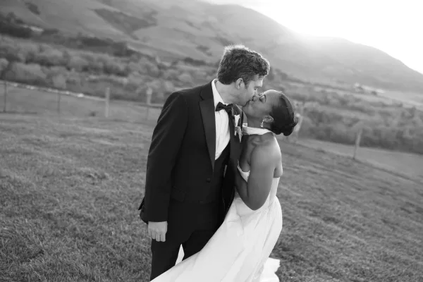Wedding kiss portrait of bride and groom in a tuxedo, leaning in as sunlight glows over a grassy mountain field with fence posts