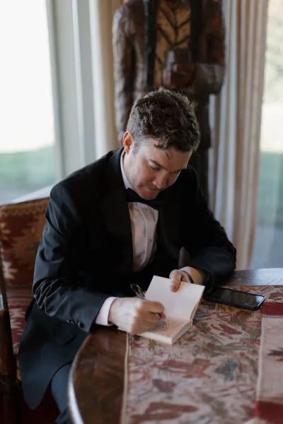 Groom portrait of groom getting ready as he writes vows in a notebook at a table, black tuxedo and bow tie in window light near greenery
