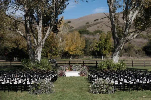 Ceremony setup with a white platform and floral ground arrangements lining the aisle, black chairs on a lawn under trees and hills
