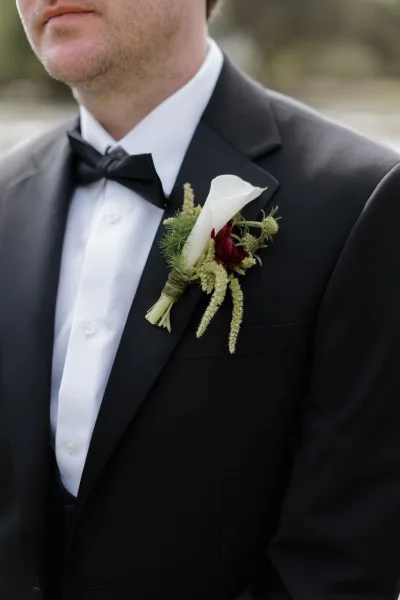 Groom boutonniere featuring a calla lily boutonniere with greenery, wrapped in ribbon on a black tuxedo lapel against an outdoor blur
