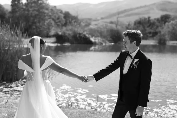 Couple portrait of bride and groom holding hands, her long veil trailing as they walk by a lakeside shoreline with mountains behind