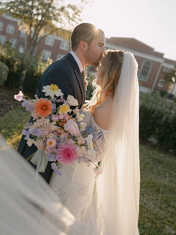 Wedding kiss portrait of bride and groom kiss, her veil and lace dress glowing in sun flare as he holds a bouquet beside a brick building