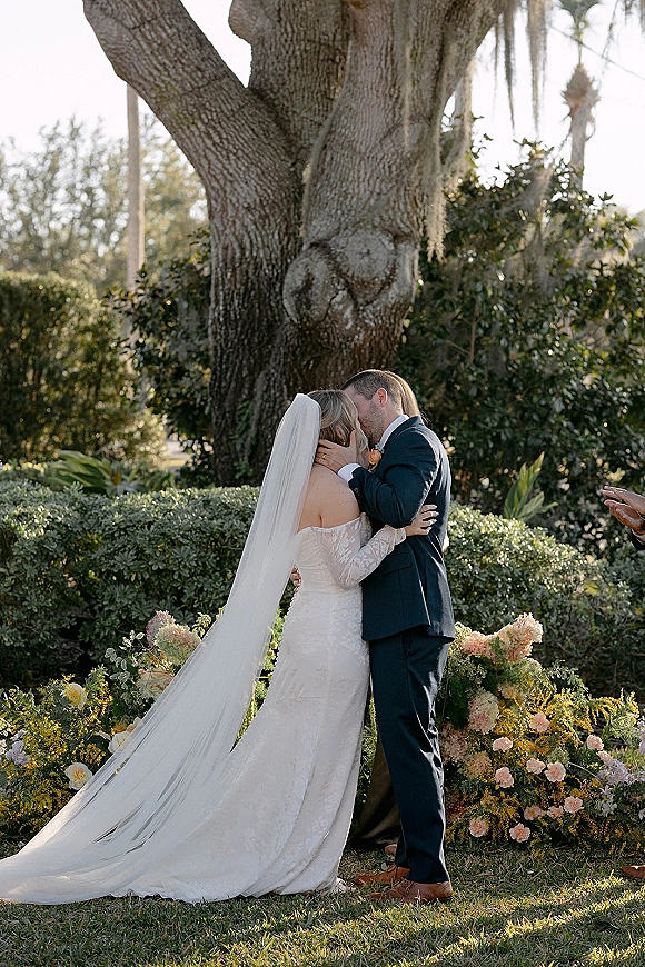 Wedding kiss during outdoor ceremony kiss as bride in long veil and lace dress embraces groom in navy suit beneath an oak tree