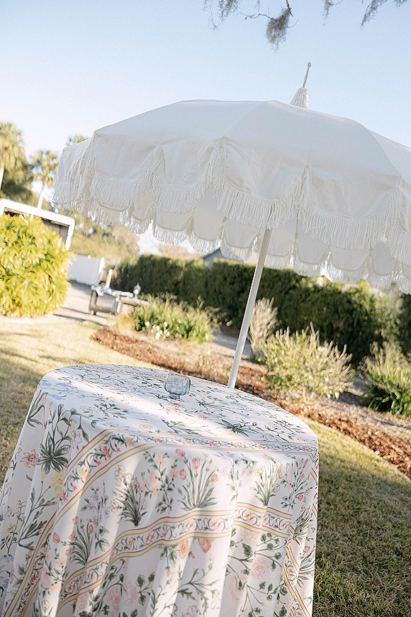 Cocktail table decor with a floral tablecloth and blue drinking glass, shaded by a white fringe umbrella on a sunny garden lawn path