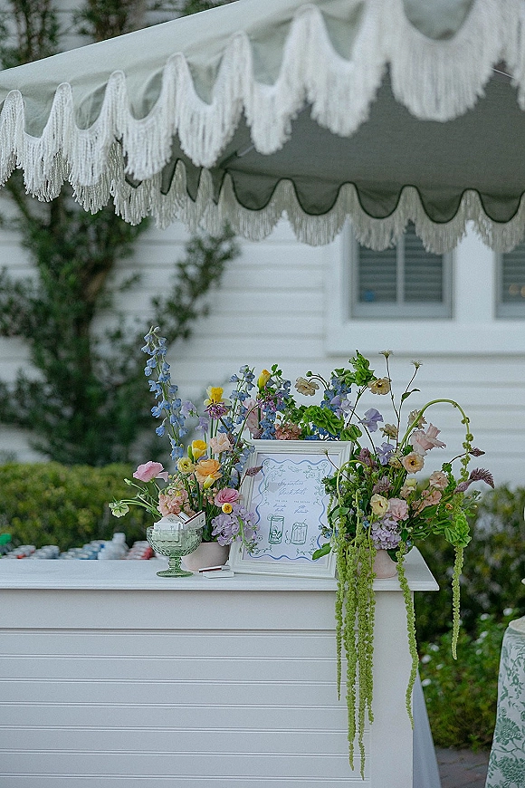 Wedding welcome table with a wedding welcome sign in a framed display, lush florals and greenery garland under a fringed canopy by a white house