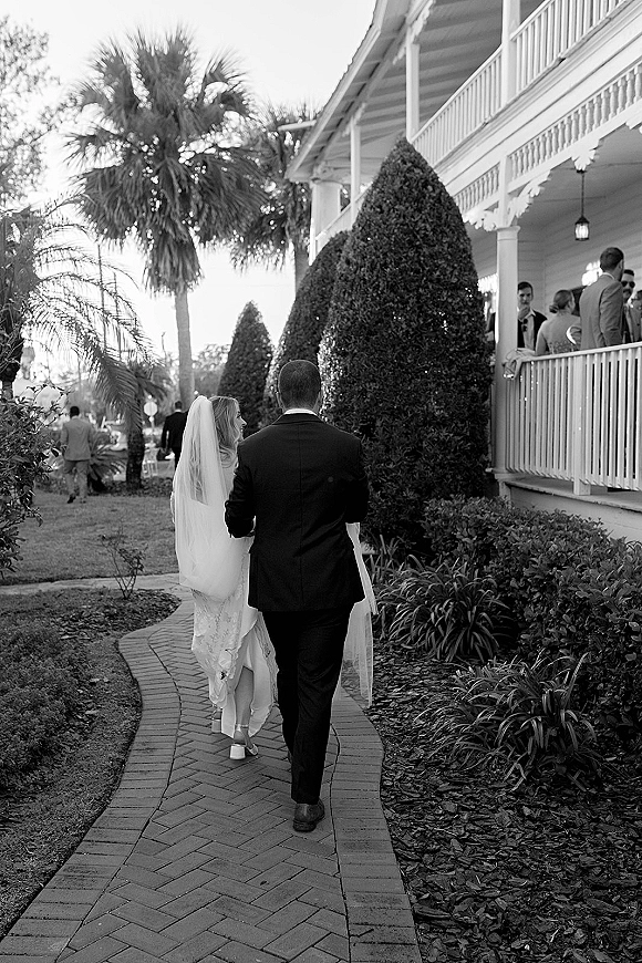 Wedding couple walking hand in hand, bride and groom from behind with long veil and boutonniere on a brick path past a porch with guests