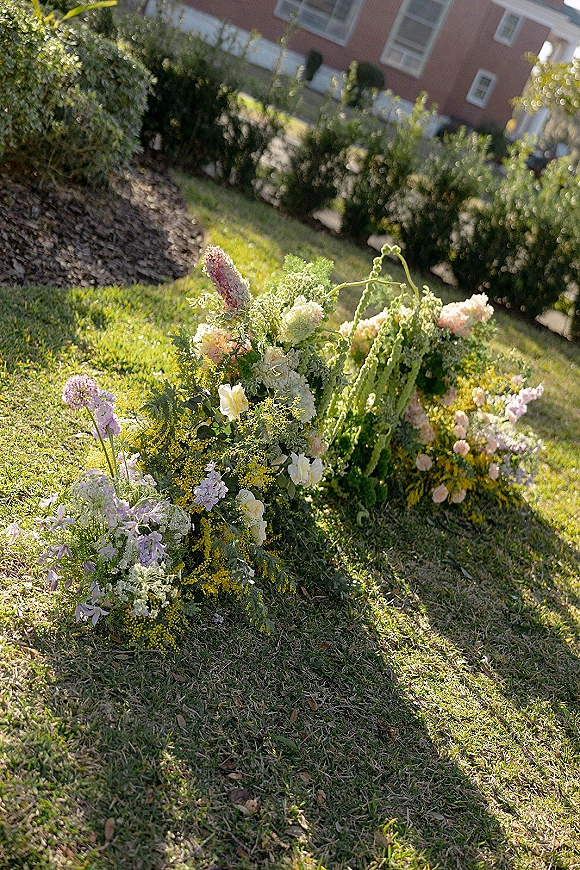 Ceremony aisle florals with grounded aisle flowers in pastel blooms and greenery lining a grass lawn walkway beside a brick building