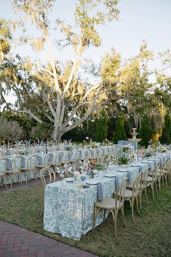 Outdoor reception tablescape with garden reception tables dressed in patterned linens, bud vase florals and taper candles under string lights by an oak tree