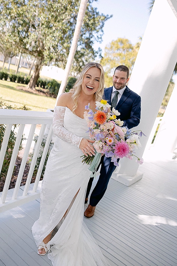 Couple portrait of bride in off shoulder lace wedding dress and groom in navy suit on a white porch, bride holding wildflower bouquet