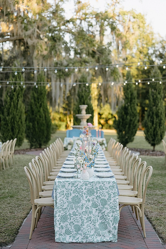 Outdoor reception table with a blue and white patterned tablecloth, wildflower centerpieces, taper candles, and string lights in a garden by a fountain