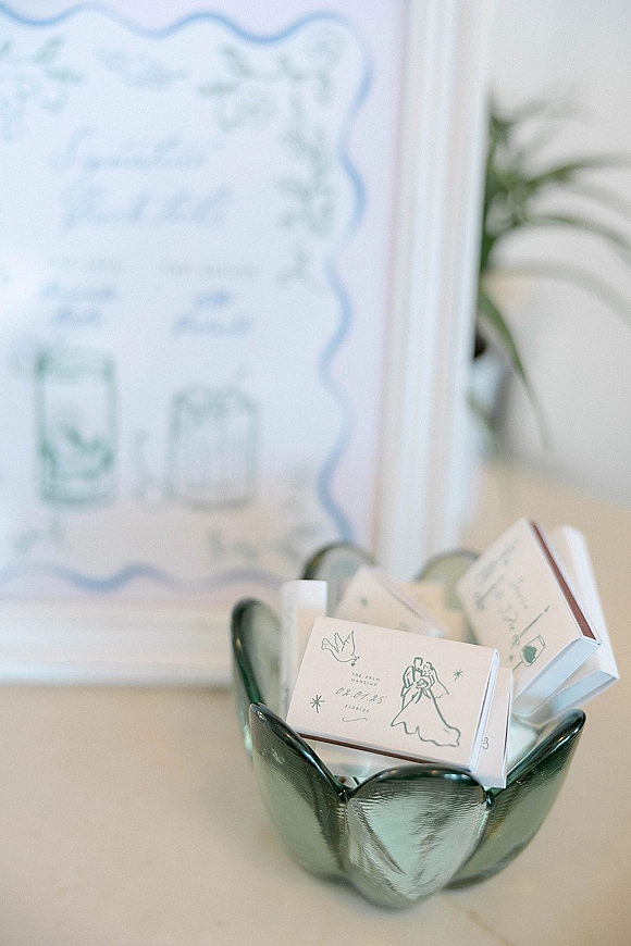 Wedding matchbooks piled in a glass bowl, showing an illustrated cover, with a blurred wedding sign and leafy tabletop in back
