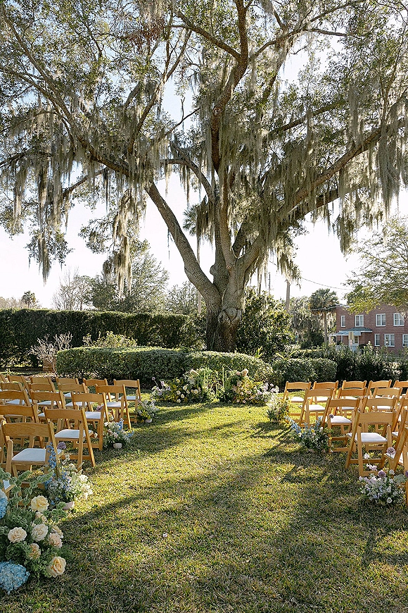 Ceremony setup with wood folding chairs in curved rows, aisle florals and petals beneath an oak tree draped in Spanish moss on a garden lawn
