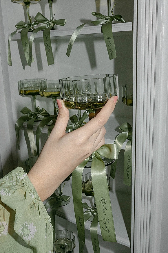 Champagne place cards on coupe glasses with satin ribbon and handwritten name tags arranged on white shelving with wall molding