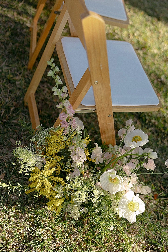 Ceremony chair decor on wood folding ceremony chairs with white cushions, wildflower and greenery arrangement beside the aisle on a sunny lawn