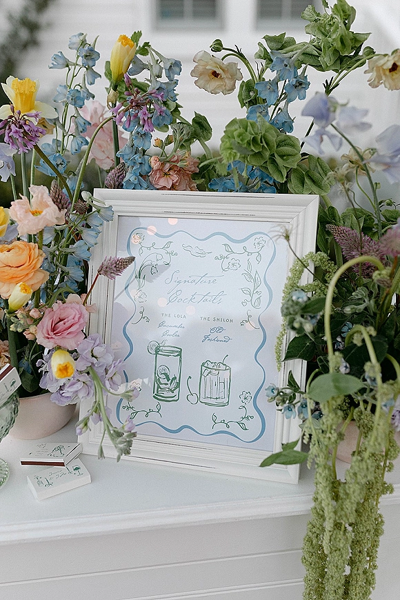 Signature cocktail sign in a white frame with illustrated drinks, pastel wildflower accents and matchboxes on a tabletop by window light