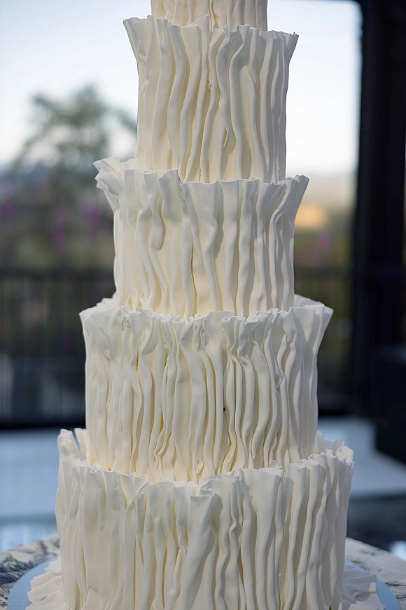 Wedding cake with white wedding cake buttercream ruffle texture on a cake stand, lit by window light with blurred greenery behind