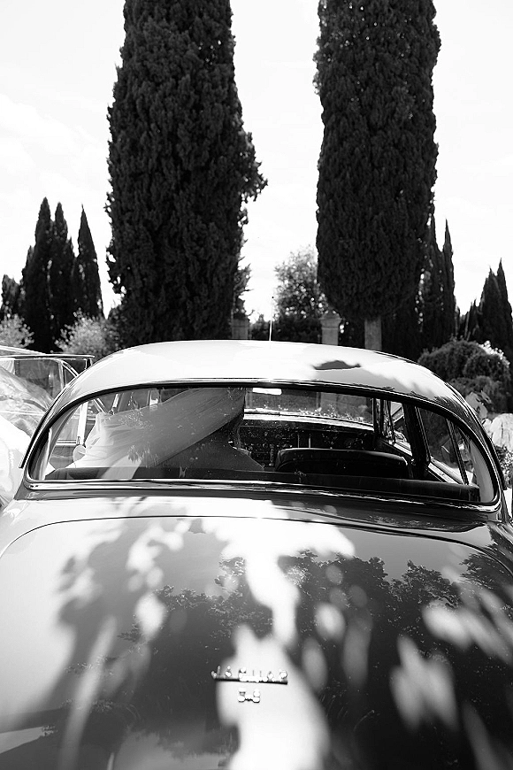 Wedding getaway car, vintage wedding car with bride’s veil draped in the window, classic emblem visible beneath tall trees and greenery