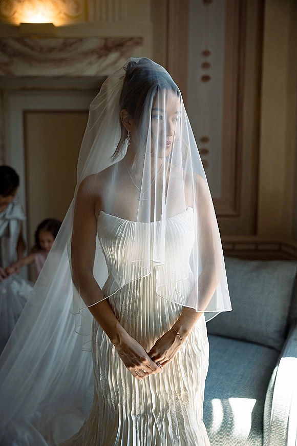 Bridal portrait of a bride in veil with sheer fabric over her face, standing by a sofa in warm window light, looking away in a strapless gown