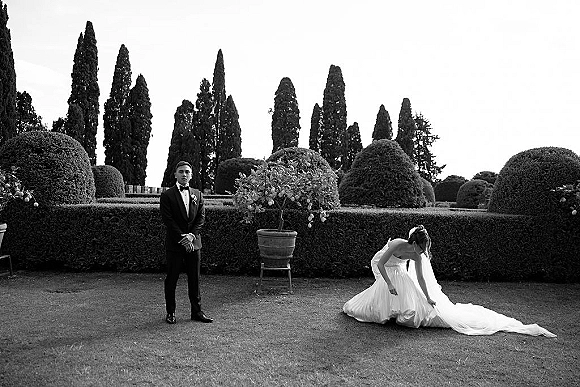 Wedding couple portrait in black and white with bride adjusting her long dress train and veil beside groom in tuxedo in a formal hedged garden