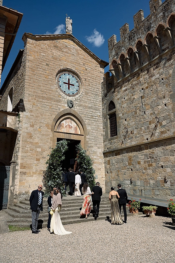 Church wedding entrance with guests walking up stone steps beneath a greenery arch, past potted flowers on a sunlit stone facade