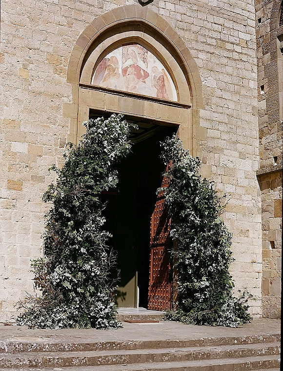 Ceremony entrance decor with eucalyptus branches framing a wooden church door, set against a stone church exterior with arched doorway and steps