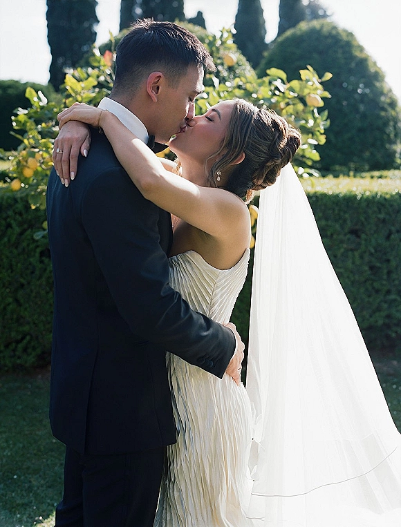 Wedding kiss as bride and groom embrace in a sunlit garden, her long veil catching the light behind a strapless dress
