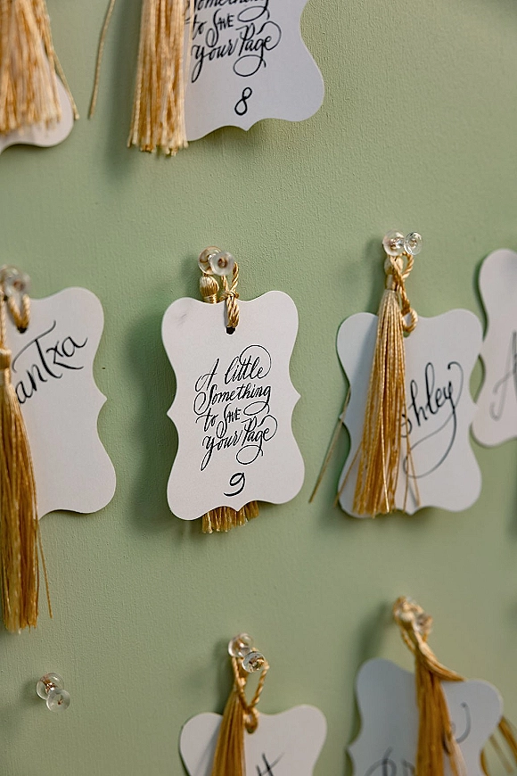 Wedding escort cards with calligraphy escort cards pinned by push pins, each with a gold tassel, displayed on a sage green board backdrop